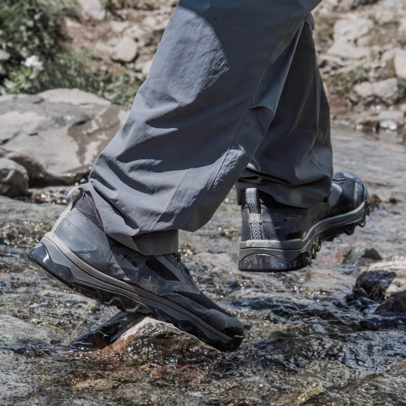 A person in gray pants crosses a small rocky stream, wearing GORUCK's Mackall shoes in Black + Forged Iron. The water is clear, revealing a few small rocks in this serene outdoor setting, reminiscent of Special Forces training grounds.