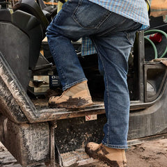 A person wearing jeans and the rugged MACV-2 Safety Boot - Mid Top by GORUCK, designed to Special Forces standards, is climbing into a heavy construction vehicle on a dirty worksite.
