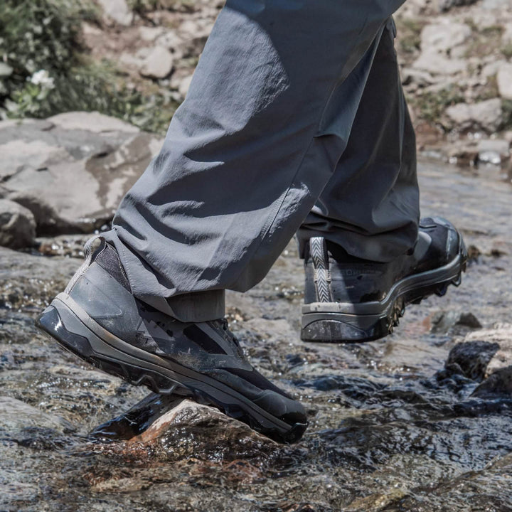 A person in gray pants crosses a small rocky stream, wearing GORUCK's Mackall shoes in Black + Forged Iron. The water is clear, revealing a few small rocks in this serene outdoor setting, reminiscent of Special Forces training grounds.
