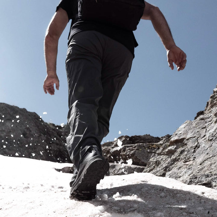 A person wearing Mackall sneakers in Black + Forged Iron by GORUCK climbs a snowy slope under a clear blue sky. Snowflakes scatter with each step forward, revealing rocky terrain in the background. The scene exudes a sense of adventure and wilderness exploration, reminiscent of Special Forces training in rugged environments.
