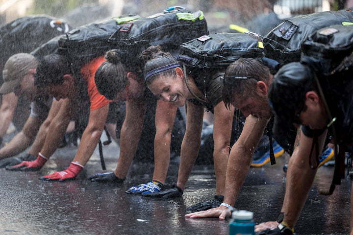 People in athletic gear do push-ups on wet ground outdoors while water sprays over them.