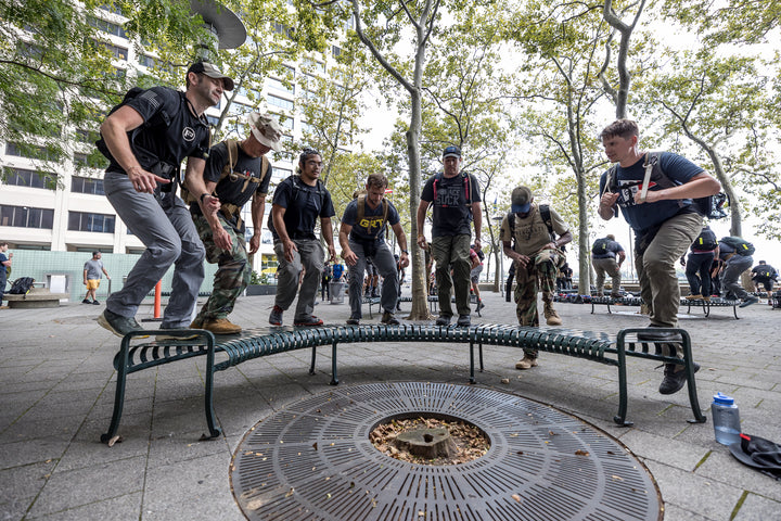 Seven people with backpacks jump over a curved metal bench in a city park with trees and tall buildings.