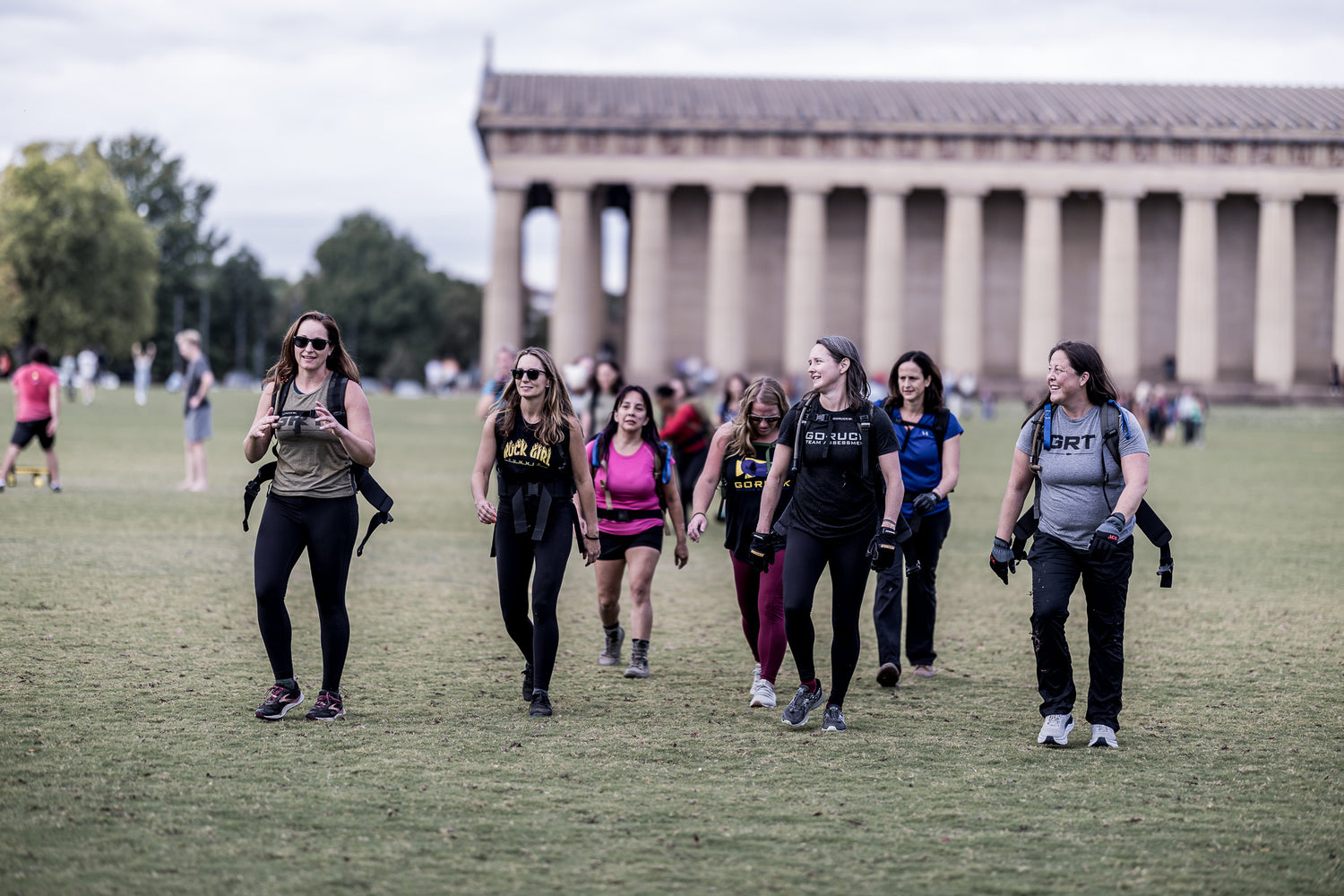 A group of women walk and smile together in front of a large building with columns on a grassy field.