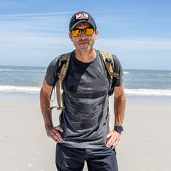Wearing a GORUCK Spearhead Tee - Tri-Blend, a man in sunglasses, cap, and backpack stands on a sandy beach with the ocean behind him.