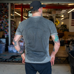 A tattooed man, seen from behind, wears a sweat-soaked gray Short Sleeve Pocket Tee - Tri-BlendX in a cluttered indoor workspace.