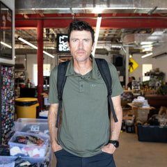 A man stands in a cluttered workspace, wearing the Men's Polo with Pocket - Tri-BlendX. Exposed pipes and shelves fill the background.