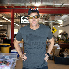 A man in sunglasses, a cap, and the GORUCK Spearhead Tee - Tri-Blend stands confidently in an industrial workshop.
