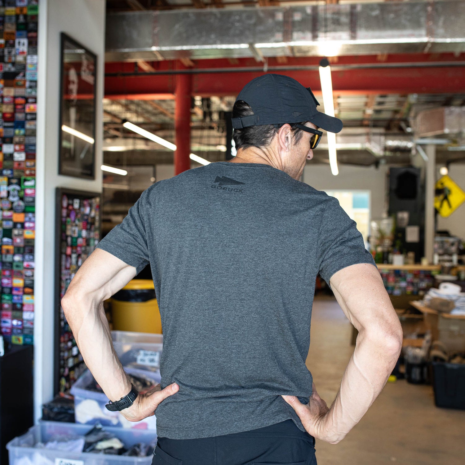 A man stands hands on hips in a cluttered office, wearing the GORUCK Spearhead Tee - Tri-Blend in gray and a black cap. The back of his shirt subtly displays the GORUCK spearhead, nodding to its Special Forces heritage.