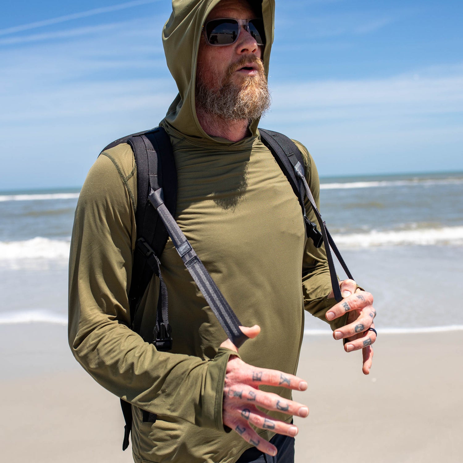 Bearded man in sunglasses and hoodie with a backpack uses T-REX Hand Straps, the ultimate rucking accessory, while strolling on a sunny beach by the ocean.