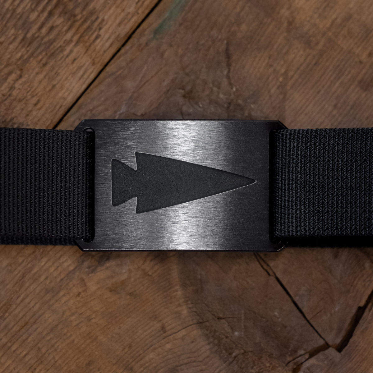 The Spearhead Nylon Webbing Belt, shown on a wooden surface, is a black belt with a matte buckle featuring an engraved arrowhead design.