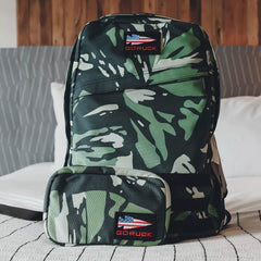 A GORUCK camouflage backpack and the GR1 Field Pocket x Gear Patrol are neatly arranged on a bed, both in Variegated Elephant Ear Camo. The backpack features the GORUCK logo alongside a stylized American flag patch. A wooden wall and white pillow serve as the backdrop, creating a display worthy of Gear Patrol.