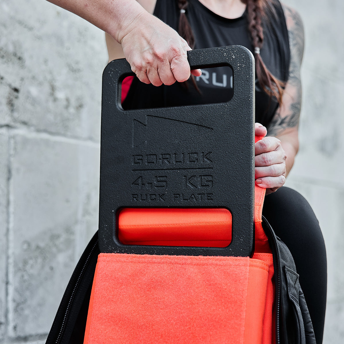 A person places a 4.5 kg Ruck Bundle into an orange-backed backpack against a concrete wall.