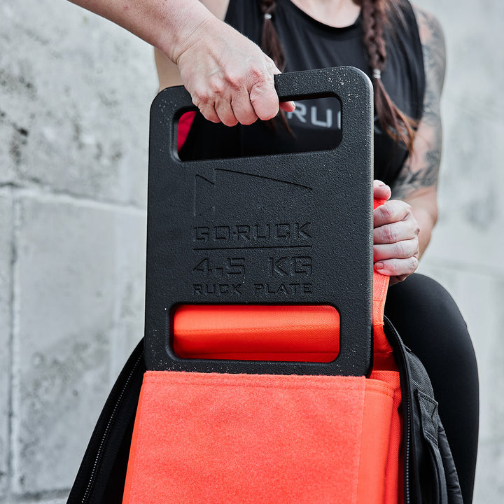 A person places a 4.5 kg Ruck Bundle into an orange-backed backpack against a concrete wall.