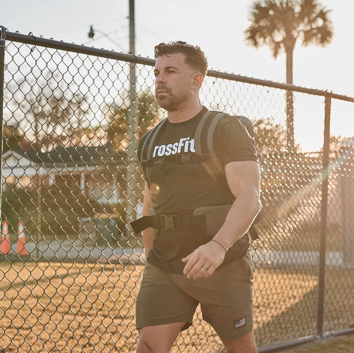 A person wearing a CrossFit t-shirt and shorts walks outdoors beside a chain-link fence, the sun setting in a warm glow. They carry a weighted backpack equipped with GORUCK's Padded Hip Belt, designed for heavy loads, ensuring comfort and stability on their journey.