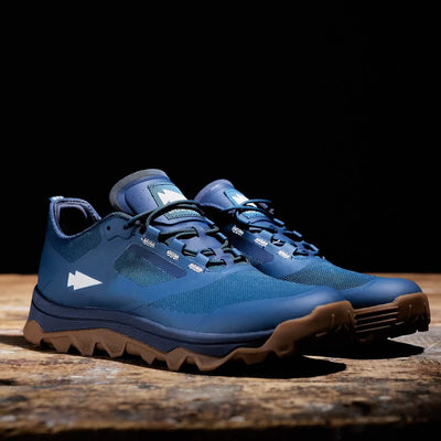 A pair of Mackall shoes in Legion Blue and Gum by GORUCK, ideal for Special Forces training, sits on a wooden surface against a dark background. The rugged design showcases breathable mesh and a prominent arrow logo on the side, combining both durability and style.