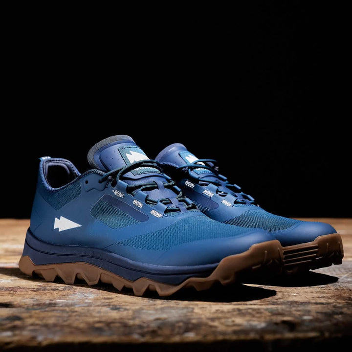 A pair of Mackall shoes in Legion Blue and Gum by GORUCK, ideal for Special Forces training, sits on a wooden surface against a dark background. The rugged design showcases breathable mesh and a prominent arrow logo on the side, combining both durability and style.