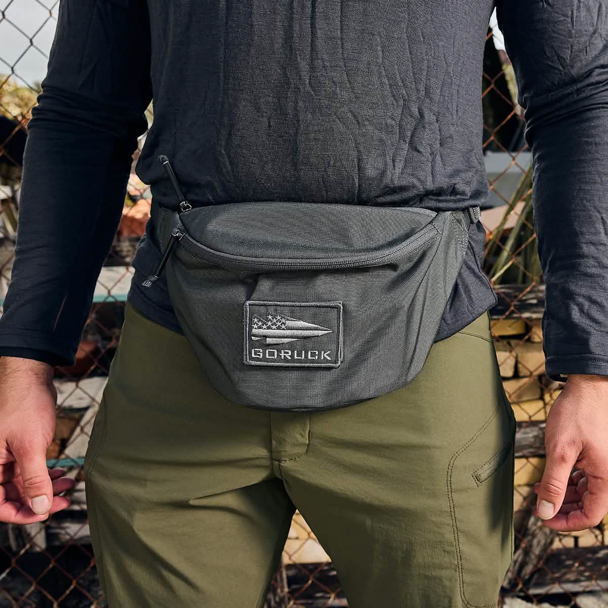 A person wears a GORUCK olive green fanny pack featuring silent zipper pulls and a GORUCK patch on the front, paired with Tactical FP1 pants in matching olive green and a black long-sleeve shirt. The background includes a fence and softly blurred outdoor elements.