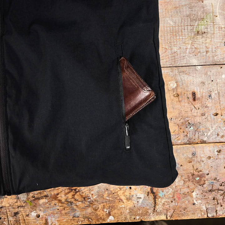 A wooden table featuring a weathered surface is partially adorned by a black GORUCK Vest of Power - ToughDry® + Fleece. Peeking out from a zipped pocket is a brown leather wallet, contributing to the rugged charm of this environmentally conscious arrangement.