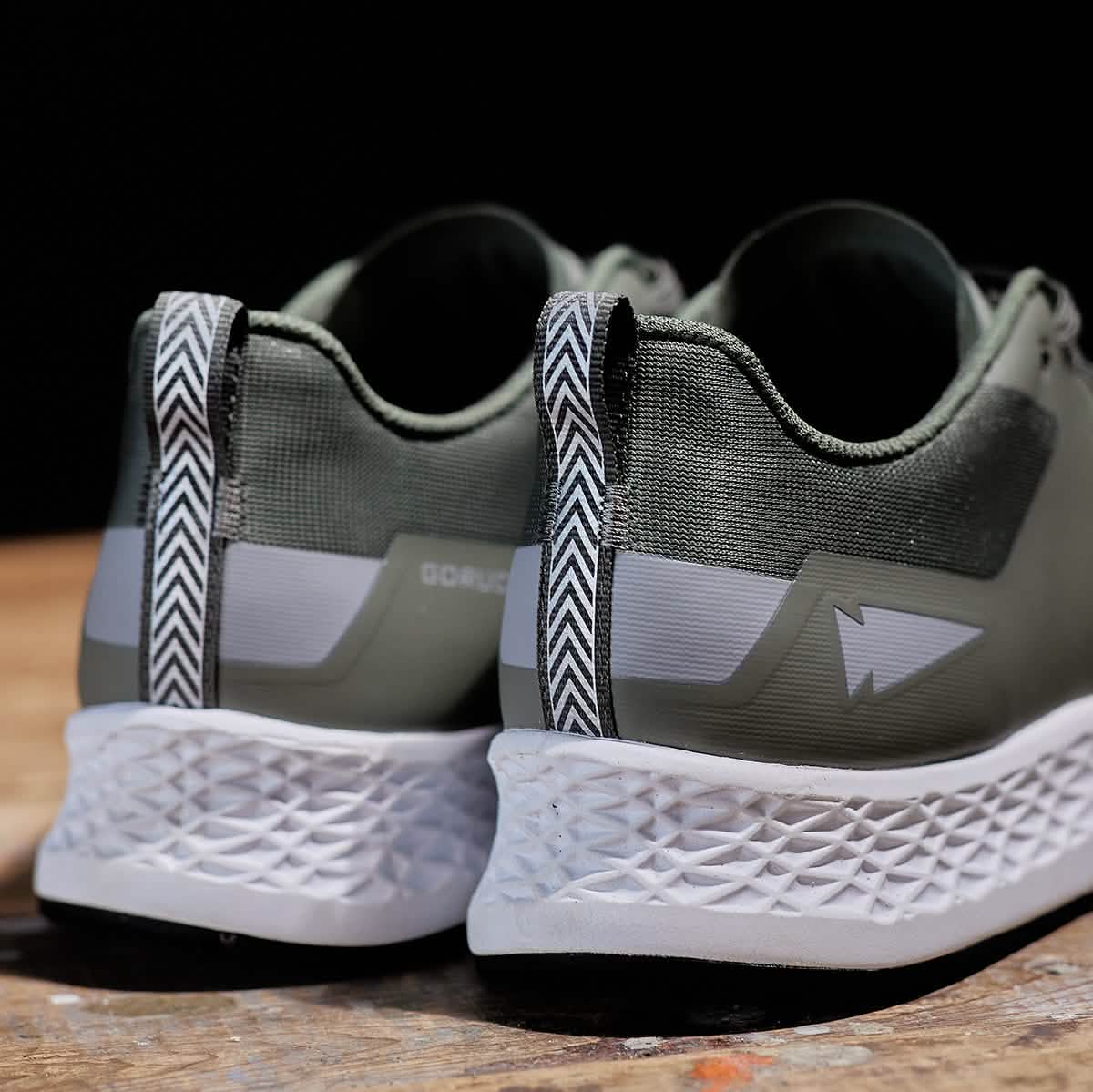 Close-up of the Women's Rough Runner - Earth green athletic shoes by GORUCK, highlighting their textured white sole and reflective details. These shoes feature an EVA midsole, chevron pattern on the heel tabs, and a geometric design on the upper. They are set on a wooden surface against a black background.