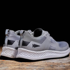 The Men's Rough Runner - Speed Grey + White athletic shoes by GORUCK showcase a geometric design in gray with black and white accents. They feature thick, textured white soles with Gradient Density support, all displayed on a wooden surface against a black background. These shoes exude the durability of Rough Runner gear when viewed from the back and side.