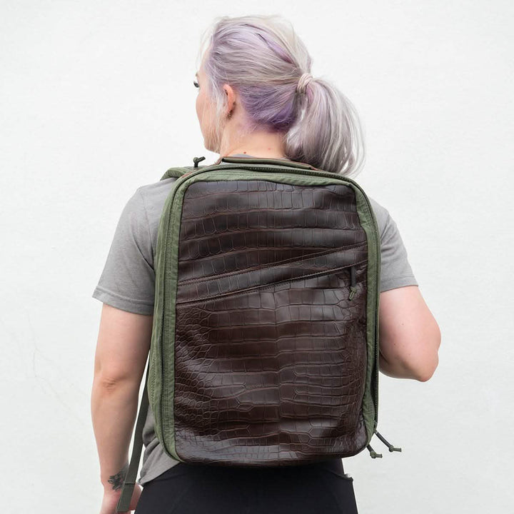 A person with light hair stands against a white background, wearing a gray shirt and showcasing the GR1 Heritage by GORUCK, which features a large green body and an alligator leather front panel.