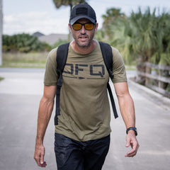 A man wearing a black baseball cap, amber-tinted sunglasses, and an olive green T-shirt over a long ruck plate carrier with a black backpack walks toward the camera.