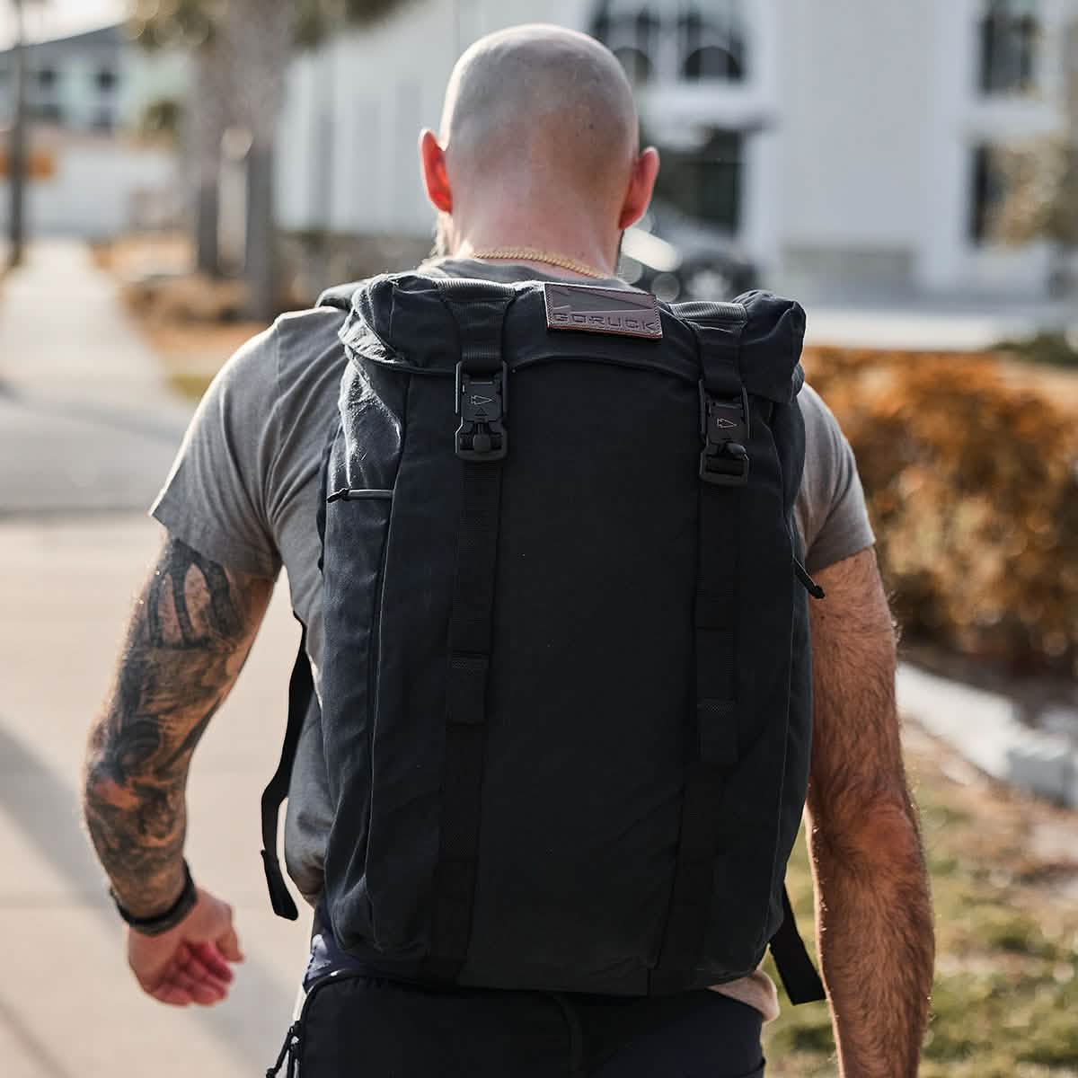 A bald man with tattoos strolls along the sidewalk, wearing a grey t-shirt and carrying a large GORUCK M23 - Waxed Canvas rucksack. The scene is set against a building backdrop, flanked by trees and a blurred street view.