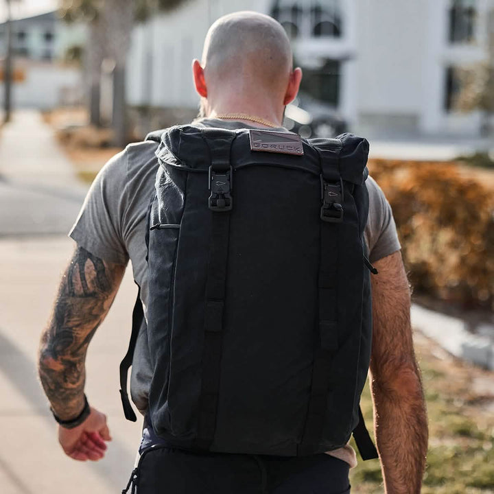 A bald man with tattoos strolls along the sidewalk, wearing a grey t-shirt and carrying a large GORUCK M23 - Waxed Canvas rucksack. The scene is set against a building backdrop, flanked by trees and a blurred street view.