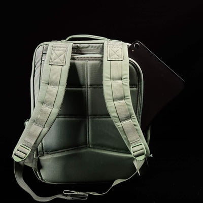 A GORUCK KR1 kids rucksack in green, featuring padded straps and visible stitching against a black background. A black tablet partially sticks out from a side pocket, making it perfect for carrying school supplies.