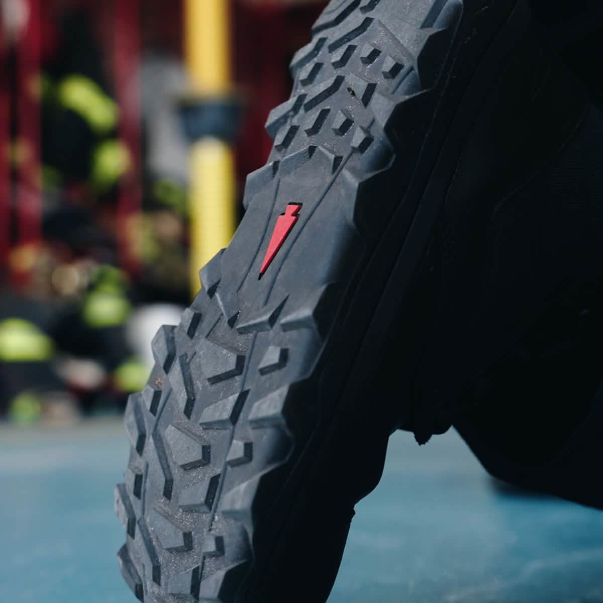 Close-up of the MACV-2 Safety Boot - High Top by GORUCK, showcasing its rugged tread and distinct red lightning bolt symbol on the sole. In the blurred background, equipment and a yellow pole suggest compliance with Special Forces standards in an industrial environment.