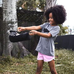 Smiling girl outdoors tossing black GORUCK sandbag in grassy yard with trees