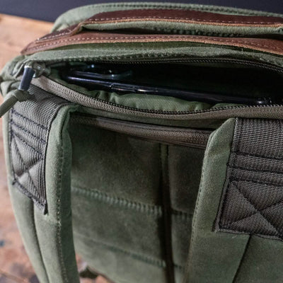 A green and brown retro canvas rucksack with a visible top zipper pocket slightly open, revealing part of a black object inside. The GORUCK GR1 Heritage backpack with an alligator leather front panel is resting on a wooden surface.