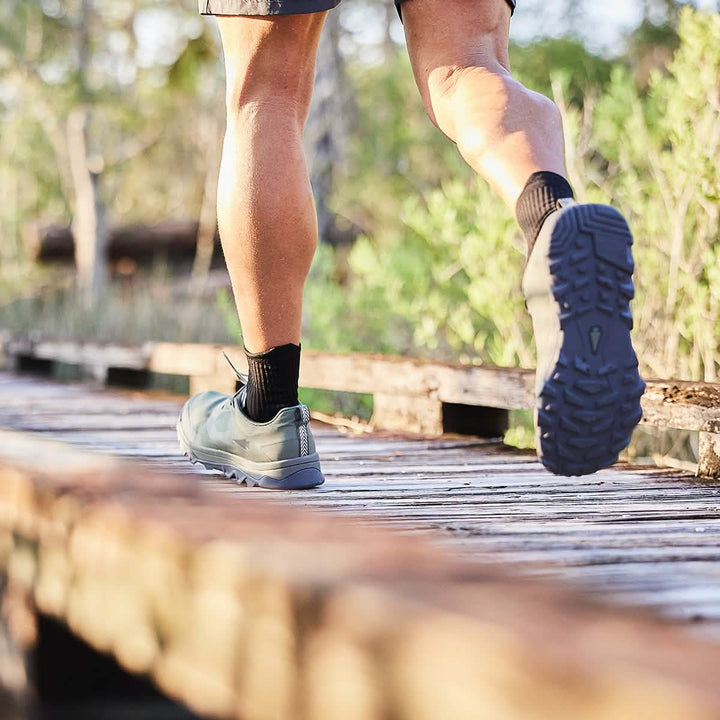 A person walking on a wooden trail in the outdoors sports Mackall - Earth + Forged Iron athletic trail shoes by GORUCK with black socks. The focus is on the legs and shoes, featuring a triple compound outsole for extra grip, against a backdrop of lush greenery and sunlight.