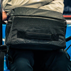 Close-up of the GR2 USA x Carryology - Ultra Black bag, made with UHMWPE fabric, resting on a person's lap while they sit on a blue seat.
