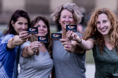Four smiling women holding GORUCK Badass Babes Basic patches outdoors, showcasing rucking community gear