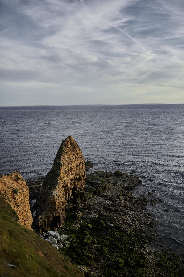 Rocky sea cliff with green moss at the water’s edge under a cloudy sky, overlooking a calm ocean.