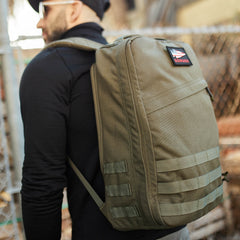 Man in black clothing and cap wears a large olive green GR1 USA - Cordura rucksack with a patch on the upper pocket.