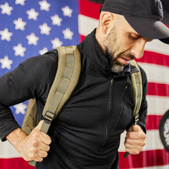 A man with a GR1 USA - Cordura rucksack stands before a large American flag, looking downward.