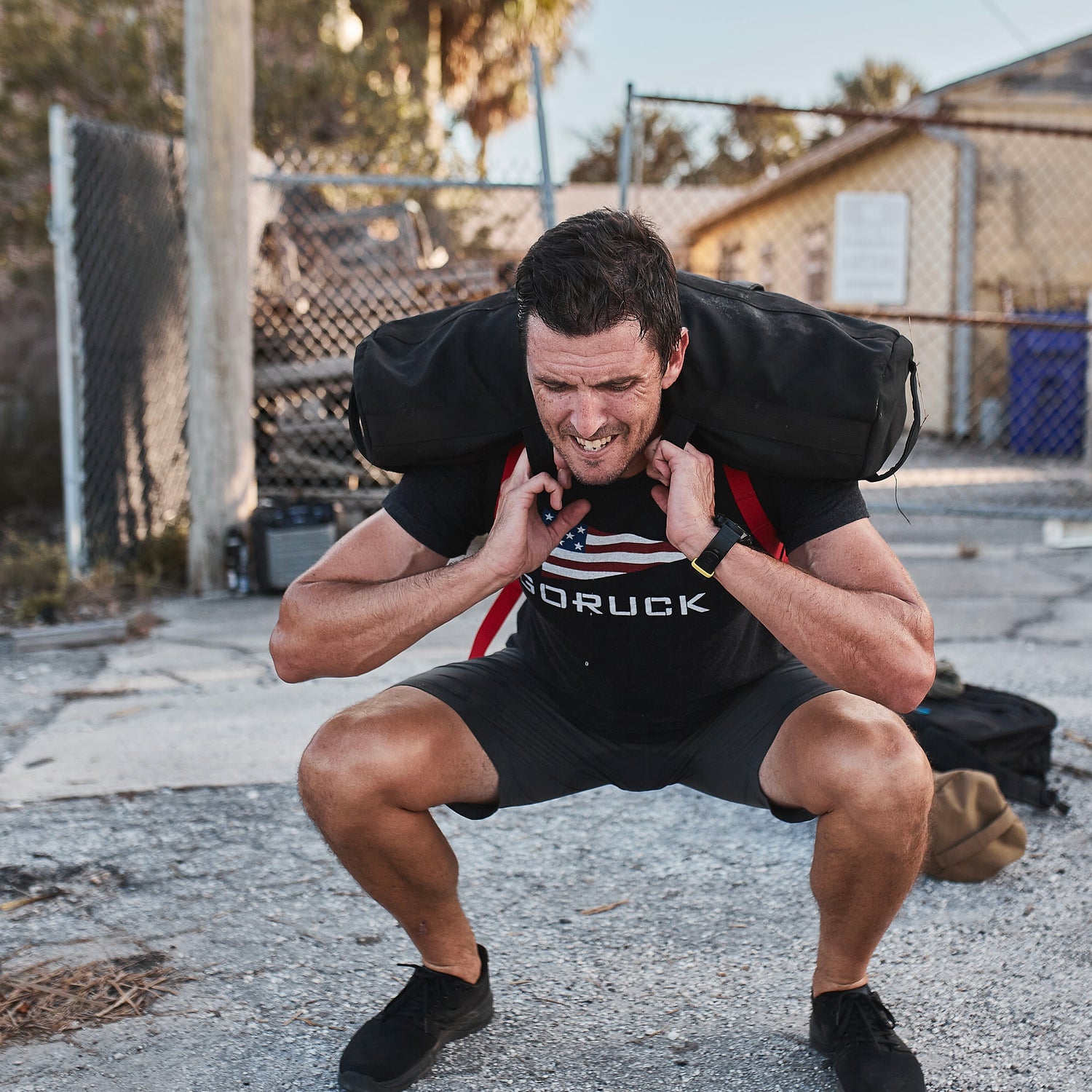 Man squats outdoors lifting a Sandbag Kit (w/ Filler Bag) on his shoulders, wearing a black GORUCK shirt—showcasing the durability and performance of training sandbags backed by a lifetime guarantee.