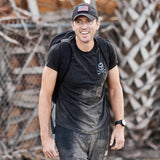 Man wearing a black cap, the GORUCK Tribe Tee - Tri-BlendX featuring a skull logo, and a backpack, smiling outdoors near a fence—ready to take on the next GORUCK Challenge as part of the real community found within GORUCK Tribe.