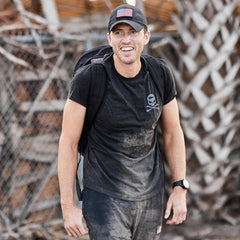 Man wearing a black cap, the GORUCK Tribe Tee - Tri-BlendX featuring a skull logo, and a backpack, smiling outdoors near a fence—ready to take on the next GORUCK Challenge as part of the real community found within GORUCK Tribe.