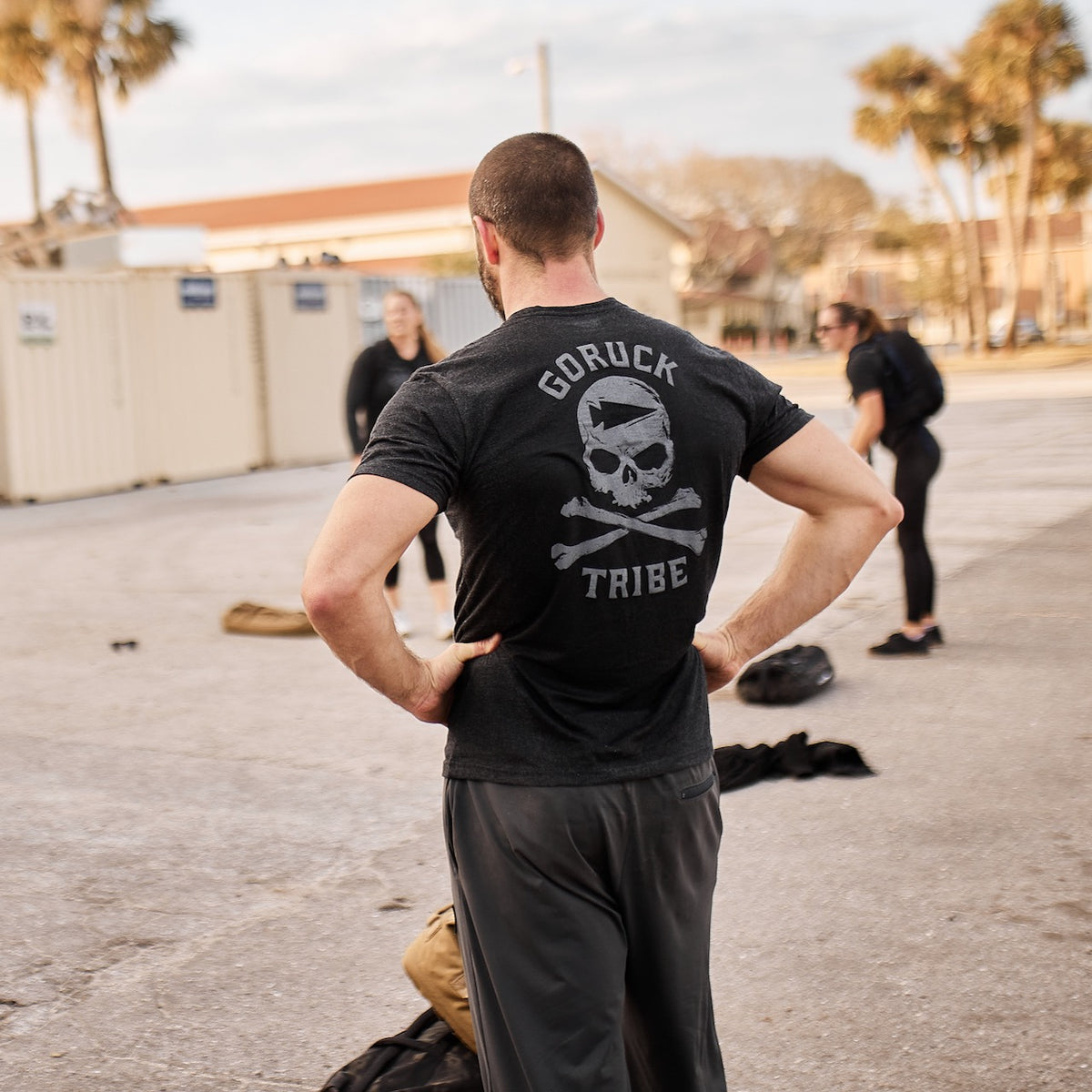 A man wearing the GORUCK Tribe Tee - Tri-BlendX stands outdoors among his community, with people and gear bags nearby—ready for the next GORUCK Challenge.