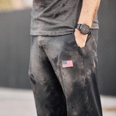 A person wearing Men’s American Training Sweatpants, featuring a tough design with an American flag patch, paired with a black watch and gray shirt visible—crafted with pride and made in the USA.