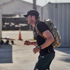 A man runs outdoors near buildings and an orange traffic cone, wearing a camo backpack, cap, and Men’s USA Training Shorts - Flag - ToughStretch.