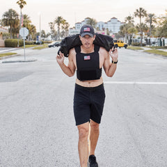 A man wearing the Training Weight Vest - Black and shorts walks down a street, holding a jacket over his shoulders.