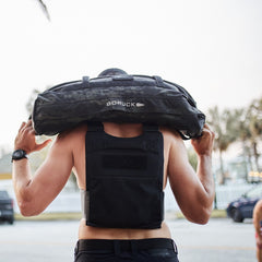 A person wearing the Training Weight Vest - Black carries a weighted sandbag on their shoulders outdoors, seen from behind.
