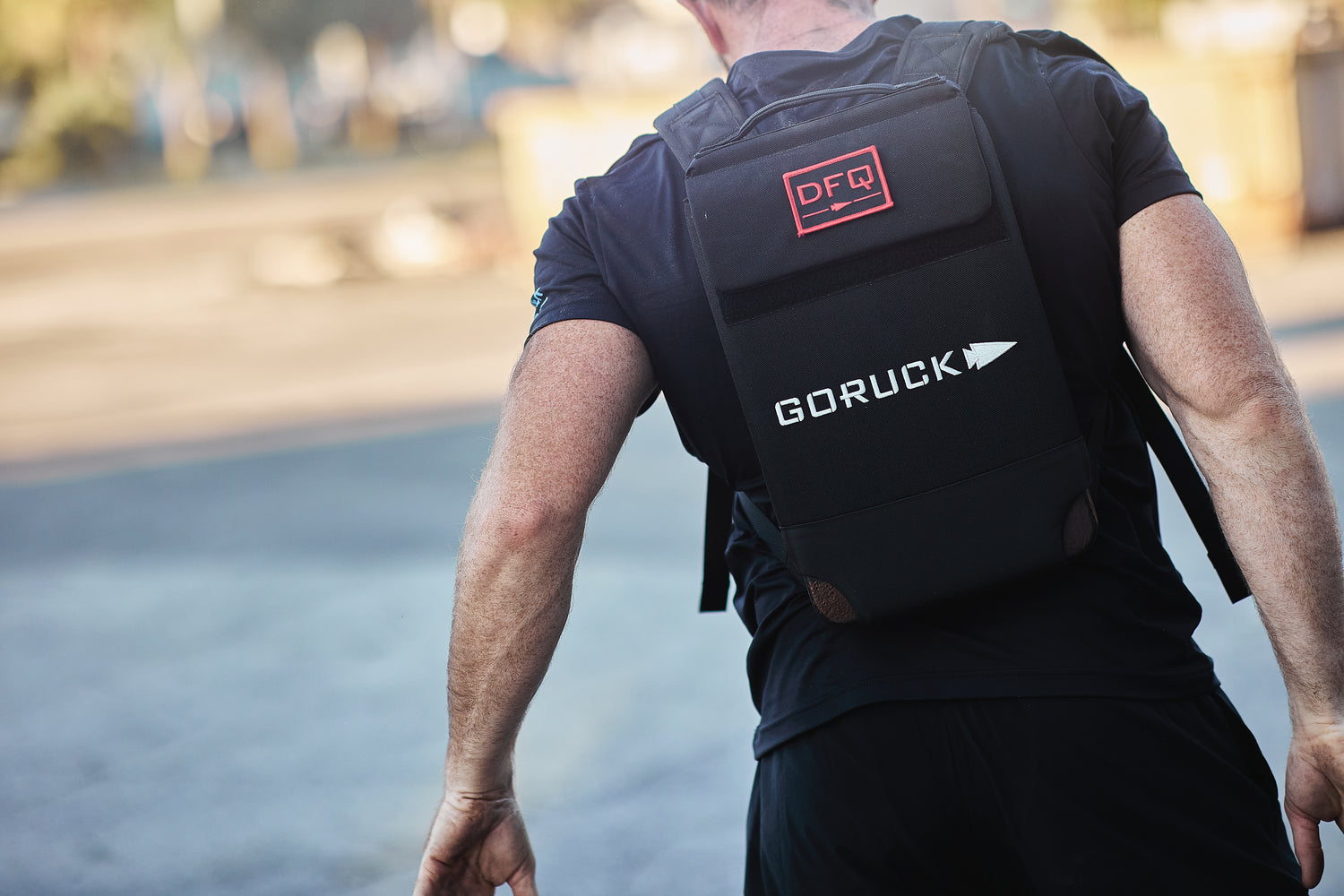 A person outdoors, seen from behind, wearing the RPC Long + 30LB (Long) Plate Bundle in black with a red DFQ patch, highlighting its ergonomic lumbar support.