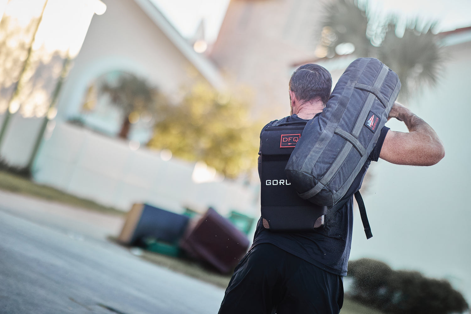 A man outdoors lifts a heavy sandbag onto his shoulder while wearing the RPC Long + 30LB (Long) Plate Bundle for weighted training.
