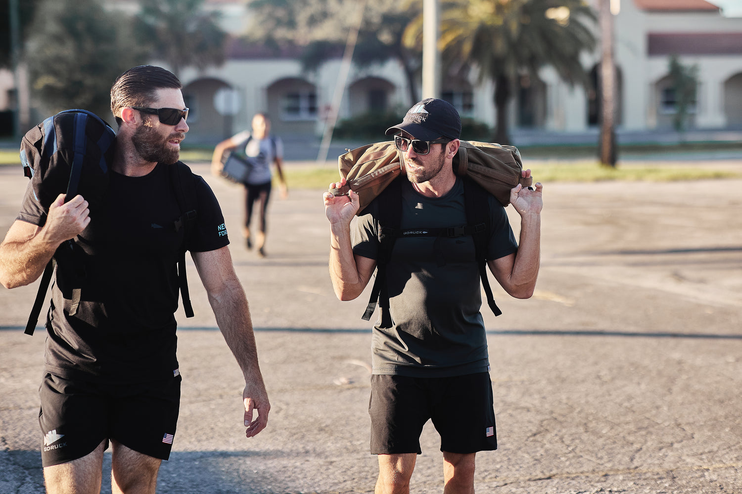 Two men in athletic wear carry weighted bags on their shoulders while walking outside in sunlight, using the RPC Long + 30LB (Long) Plate Bundle for ergonomic lumbar support.