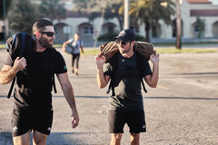 Two men in athletic wear carry weighted bags on their shoulders while walking outside in sunlight, using the RPC Long + 30LB (Long) Plate Bundle for ergonomic lumbar support.